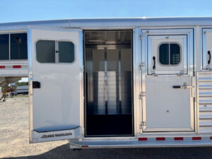 A close-up view of an open door on a shiny aluminum Elite 4 horse trailer, revealing the empty interior. The exterior features windows, vents, and polished metal panels. The trailer is parked on a gravel lot under a clear sky. -Stillwater Trailer Sales