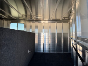 Interior of an empty metal trailer with a window on the left, padded divider, carpeted floor, and a rail mounted to the right wall. The reflective metal panels evoke GRACIA’s sleek style—perfect for transporting your Horse Mustang. -Stillwater Trailer Sales