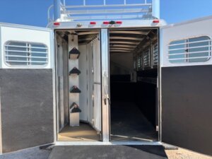 The image shows the open rear doors of a GRACIA Elite 4 horse trailer, revealing saddle racks on the left and an empty stall area inside. Bright sunlight highlights the white and black exterior and metal bars on the windows. -Stillwater Trailer Sales