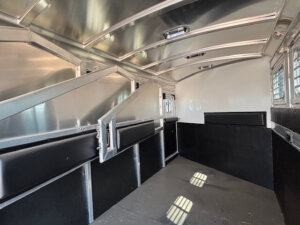 Interior view of an empty GRACIA Elite 4 horse trailer with padded dividers and metal walls; natural light enters through slatted windows, casting shadows on the floor—perfect for your Horse Mustang’s comfort. -Stillwater Trailer Sales