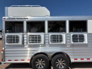 A silver Elite 4 Horse trailer with three side windows, each with ventilation bars. The trailer has dual axles with alloy wheels and visible Elite Trailers branding. The sky is clear and blue—a perfect day for your Mustang or GRACIA to travel in style. -Stillwater Trailer Sales