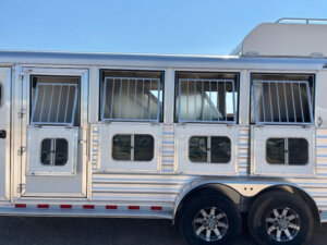 Silver Elite 4 Horse trailer with three side-by-side stalls, each featuring barred windows and hinged access doors. The trailer is parked on gravel and has two visible wheels with alloy rims. The sky is clear. -Stillwater Trailer Sales