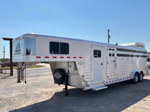An Elite 26 Stock Combo silver gooseneck livestock trailer is parked on gravel. The trailer features multiple side vents, a black spare tire cover, tandem rear axles, and a clear blue sky with power lines in the background. -Stillwater Trailer Sales