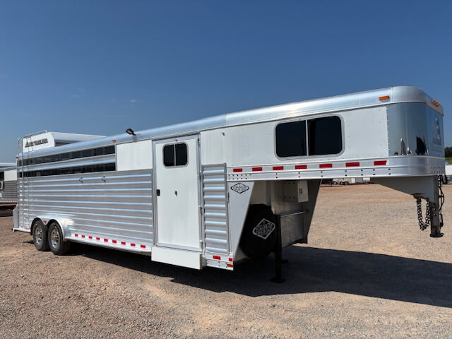 An Elite 26 Stock Combo large silver gooseneck livestock trailer with multiple windows and a side door is parked on a gravel lot under a clear blue sky. -Stillwater Trailer Sales