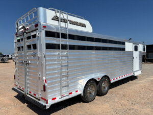 An Elite 26 Stock Combo Trailer with a corrugated metal exterior, ventilation slats, tandem axles, and rear loading doors is parked on a gravel lot under a clear blue sky. -Stillwater Trailer Sales