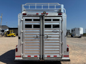 A rear view of a silver Elite 26 Stock Combo livestock trailer with closed double doors and ventilation slats, parked on a gravel lot. Other vehicles and trailers are visible in the background under a clear blue sky. -Stillwater Trailer Sales