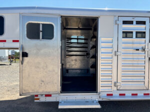 Open side door of an Elite 26 Stock Combo horse trailer reveals interior with saddle racks mounted on the left wall. Sunlight is visible outside on gravel ground. The trailer has horizontal vents and a step at the entrance. -Stillwater Trailer Sales