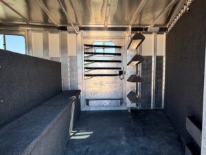 Interior of the Elite 26 Stock Combo horse trailer with carpeted bench seating on the left, metal wall and door in the center, and saddle racks with black padding on the right. Sunlight streams through windows on both sides. -Stillwater Trailer Sales
