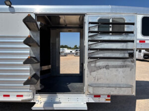 An Elite 26 Stock Combo metal horse trailer with an open side door reveals multiple saddle racks on the door and inside wall, set against a sunny outdoor background with gravel and vehicles visible. -Stillwater Trailer Sales
