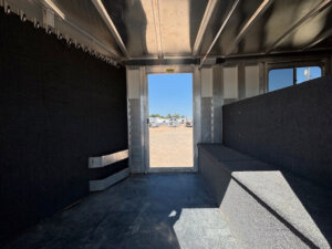 Interior of an empty Elite 26 Stock Combo trailer, featuring black padded walls and benches. Sunlight streams through the open door, offering a glimpse of vehicles parked outside. -Stillwater Trailer Sales