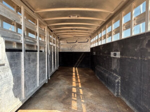 Interior view of an empty Elite 26 Stock Combo trailer with metal walls, ribbed ceiling, small windows along the top sides, and a textured brown floor. Sunlight casts shadows through the windows onto the floor. -Stillwater Trailer Sales