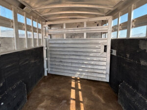 Interior of an empty Elite 26 Stock Combo trailer with metal walls and a grated partition gate in the middle. Sunlight creates shadows on the brown floor, and ventilation slats line the sides for airflow. -Stillwater Trailer Sales