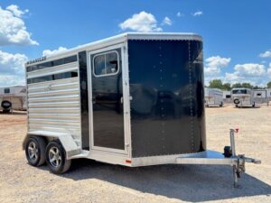 A two-axle, black and silver enclosed livestock trailer with a side door and small window is parked on a gravel lot under a mostly sunny sky, with several other trailers in the background. -Stillwater Trailer Sales