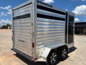 A silver Wrangler-branded horse trailer with dual axles and black accents is parked on gravel under a blue sky with scattered clouds. The trailer has a rear door and side window. -Stillwater Trailer Sales