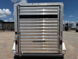 A silver, metal livestock trailer with the word WRANGLER on the back door is parked on gravel under a partly cloudy sky. The rear door is closed and ribbed in design for added strength. -Stillwater Trailer Sales
