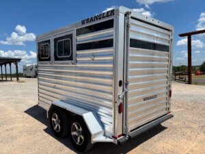 A silver, metal horse trailer with two wheels and tinted windows is parked on a gravel surface under a blue sky with scattered clouds. The word WRANGLER is printed on the side and rear of the trailer. -Stillwater Trailer Sales