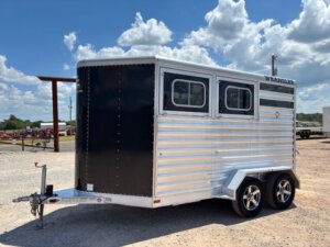 A silver and black two-horse trailer with two windows and the word WRANGLER on the side, parked on gravel under a partly cloudy sky. -Stillwater Trailer Sales