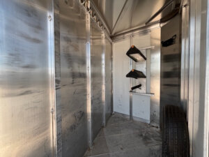 Interior of a metal shed with reflective walls, three black shelves with wooden brackets on the far wall, a mounted bracket, and a spare tire on the floor to the right. The floor appears to be concrete. -Stillwater Trailer Sales