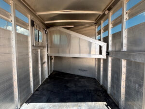 Interior of an empty metal livestock trailer with slatted sides, a black rubber floor, and a diagonal partition gate near the front. Sunlight streams in through the side vents. -Stillwater Trailer Sales