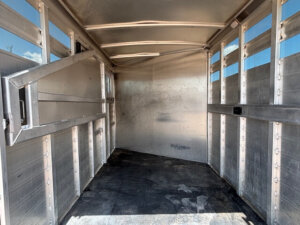 Interior view of an empty metal livestock trailer with a divider gate open to the left and a black rubber floor mat. Sunlight filters in through the slats along the sides and roof. -Stillwater Trailer Sales