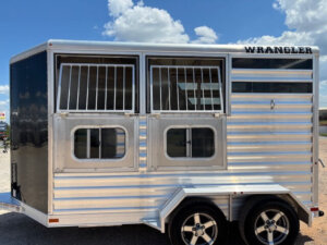 A silver Wrangler horse trailer with barred windows and a black rear section is parked outside on gravel, under a partly cloudy sky. The trailer has two axles and modern wheels. -Stillwater Trailer Sales