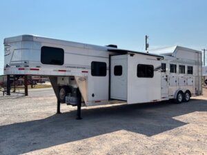 A large white gooseneck horse trailer, labeled Laramie Edition, is parked on gravel. This Living Quarters custom trailer, offered by horse and livestock trailer sales, features multiple windows, two axles, and an extended side slide-out section. -Stillwater Trailer Sales