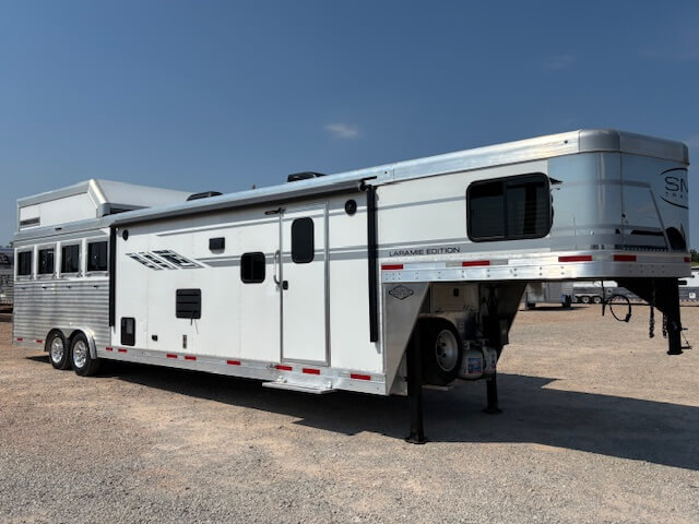 A large silver and white gooseneck horse and livestock trailer with living quarters is parked on gravel under a clear blue sky. The trailer features several windows and ventilation openings on the side, ideal for horse and livestock trailer sales. -Stillwater Trailer Sales