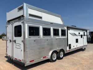 A silver and white horse trailer with multiple windows, two axles, and an attached living quarters, parked on a gravel lot under a clear blue sky—perfect for those seeking quality new and used trailers or expert horse and livestock trailer sales. -Stillwater Trailer Sales