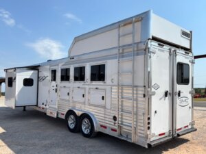 A large white and silver horse trailer with multiple windows and storage compartments, two axles with shiny wheels, a side ladder, and a slide-out section—perfect for horse and livestock trailer sales—parked on a gravel surface under a clear sky. -Stillwater Trailer Sales