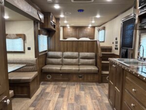 Interior of an RV with wood-paneled walls and flooring, a brown leather sofa, dining booth, kitchen area with a sink, and stairs leading up to a bed area—perfect for those seeking Living Quarters custom options found in luxury Custom Show Trailers. -Stillwater Trailer Sales