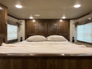 A neatly made bed with white pillows and a blanket anchors this Living Quarters custom wooden-paneled room, featuring closed cabinets above, windows on either side with light curtains, and ceiling lights aglow. -Stillwater Trailer Sales