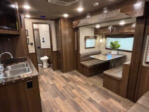 Interior of an RV with wood-paneled walls and floors, a kitchen area with a sink, built-in seating around a dining table, and windows with blinds—perfect inspiration for Living Quarters custom in new and used trailers or horse and livestock trailer sales. -Stillwater Trailer Sales