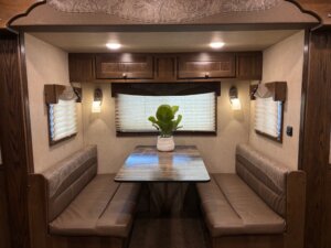 A dining nook inside an RV with two brown cushioned benches, a wooden table, overhead cabinets, wall-mounted lights, and a potted plant centerpiece. Perfect for relaxing on your next adventure from new and used trailers or custom show trailers. Three windows with blinds are visible. -Stillwater Trailer Sales