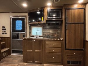 A small RV kitchen features brown cabinets, a sink, a wall-mounted TV, a microwave, refrigerator, and stovetop. To the left are stairs and a door with a window—perfect for new and used trailers or horse and livestock trailer sales. -Stillwater Trailer Sales