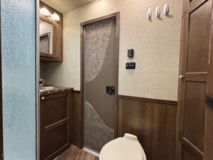 A small bathroom in one of our Living Quarters custom trailers features a toilet, brown patterned door, sink with mirror, wood-paneled walls, cabinet, control panels, hooks, and part of a frosted glass shower. -Stillwater Trailer Sales