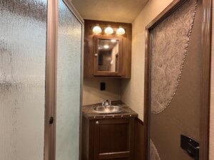 A small bathroom with a glass shower door on the left, a wood vanity with a sink and storage below, and a mirrored medicine cabinet above features Living Quarters custom touches—a perfect fit for new and used trailers from leading trailer sales. -Stillwater Trailer Sales
