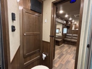 View from a bathroom looking into the main living area of an RV from Custom Show Trailers. The bathroom has a wooden door, toilet paper holder, and control panels; the living area features wood floors, cabinets, and a brown sofa. -Stillwater Trailer Sales