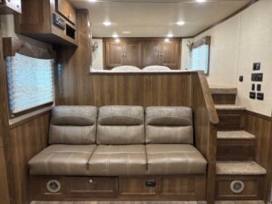 A tan leather sofa sits in front of a wooden wall with built-in cabinets above. Carpeted stairs on the right lead up to a raised bed, showcasing the comfort and style of Living Quarters custom designs found in new and used trailers. -Stillwater Trailer Sales