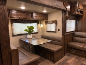 A dining area inside an RV with a wooden table, cushioned benches, wood-paneled walls and cabinets, two windows with blinds, and a potted plant on the table—showcasing the comfort found in Living Quarters custom trailers. The floor is wood and carpeted. -Stillwater Trailer Sales