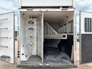 The image shows the open back doors of a Custom Show Trailer. Inside, there are saddle racks on the left and a partitioned area for transporting horses on the right, with barred windows for ventilation. -Stillwater Trailer Sales