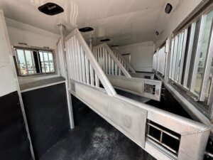 Interior view of a horse trailer with partitioned stalls, metal bars, and side windows allowing light in. The black floor and white walls with black padding near the bottom reflect the quality found in our new and used trailers. -Stillwater Trailer Sales