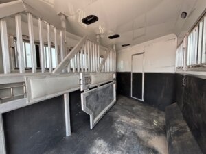 Interior of an empty horse trailer with metal dividers and padded walls, featuring barred windows for ventilation and a closed door at the far end. The floor is black and shows signs of use—ideal for those seeking new and used trailers or Custom Show Trailers. -Stillwater Trailer Sales
