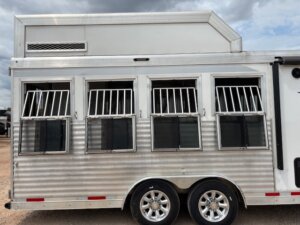 A silver Custom Show Trailer with four barred windows and black mesh screens is parked on gravel under a cloudy sky. Two chrome wheels are visible, showcasing the quality youd expect from leading trailer sales. -Stillwater Trailer Sales