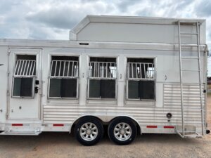 A silver, multi-compartment stock trailer with four barred windows, two tires, and a vertical ladder at the rear, parked on a gravel surface under a cloudy sky—ideal for horse and livestock trailer sales. -Stillwater Trailer Sales