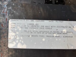 A close-up of a trailer identification label shows manufacturer SMC Trailers, date 01/2022, and specs including GVWR 7267 kg (16000 lb), model SL8414SSR. The slightly worn label hints at its Living Quarters custom design from top trailer sales. -Stillwater Trailer Sales