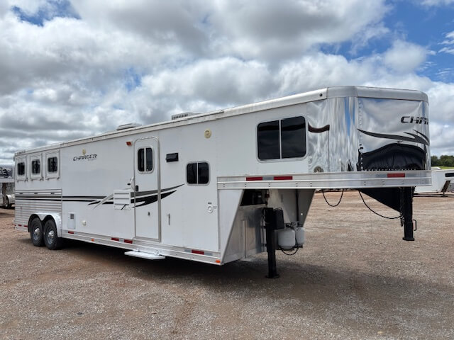 A large white fifth-wheel horse trailer with multiple windows and black detailing, from our new and used trailers selection, is parked on a gravel lot under a partly cloudy sky. -Stillwater Trailer Sales