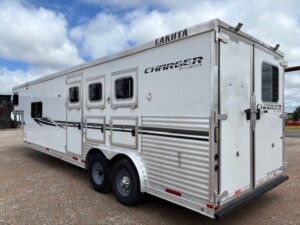 A large white Lakota Charger horse trailer with living quarters custom features and three axles is parked on gravel under a cloudy sky. The trailer has several windows and doors, with black and silver detailing on the side. -Stillwater Trailer Sales