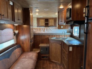 Interior of a camper with wood cabinetry, a brown sofa on the left, a compact kitchen with sink and microwave on the right, and a raised sleeping area in the back. Discover Living Quarters custom options at our trailer sales for new and used trailers. -Stillwater Trailer Sales