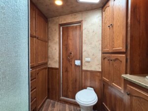A small bathroom in a custom show trailer features wood-paneled cabinets and walls, a white toilet, a door with a circular towel ring, and a textured glass shower door partially visible on the left. The floor has a wood-patterned design. -Stillwater Trailer Sales