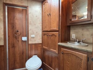 A small bathroom with wood-paneled cabinets, a toilet, wall-mounted sink with mirror above, and a door with towel ring—perfect for Custom Show Trailers or Living Quarters custom builds. The walls feature a beige and wood design. -Stillwater Trailer Sales